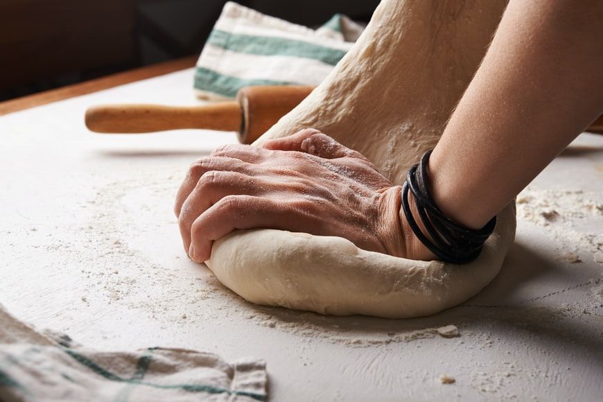 Two hands pressing pizza dough into a floured work surface beside a rolling pin.